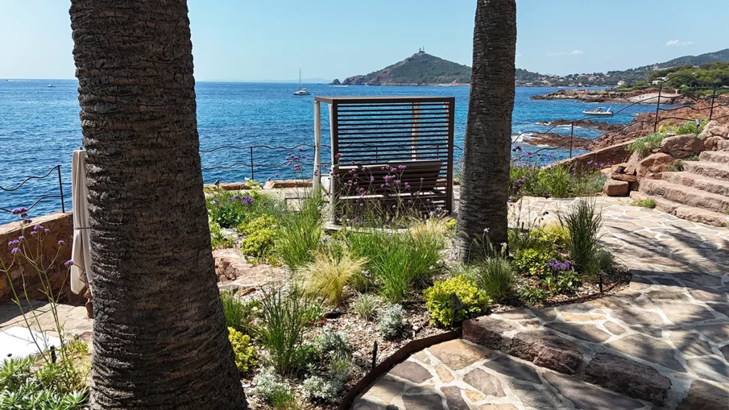 Terrasse paysagée en bord de mer avec banc en bois, marches en pierre, palmiers et végétation ornementale