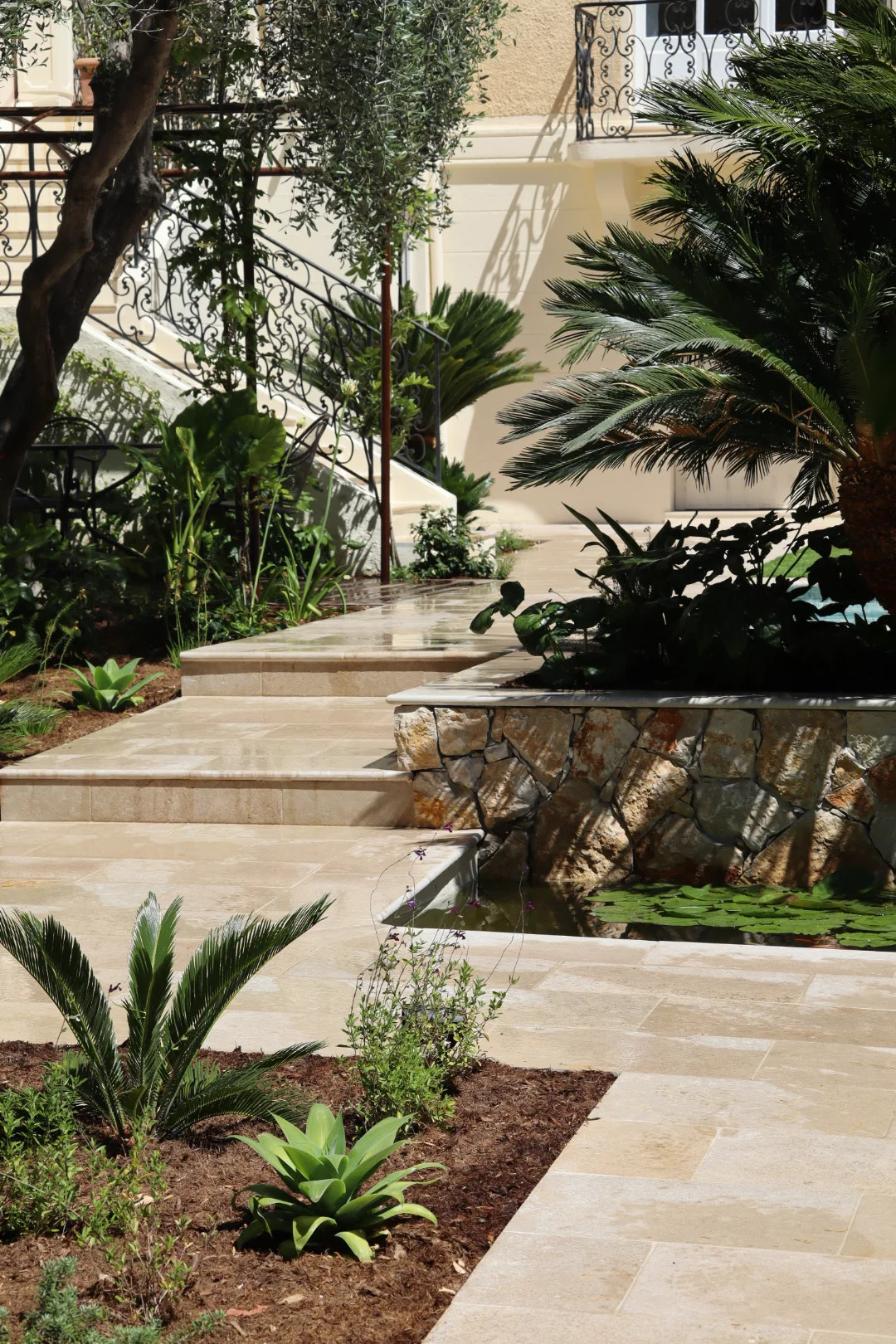 Cour de jardin avec marches et cheminements pavés, jardinières en pierre surélevées, végétation et garde-corps métalliques.
